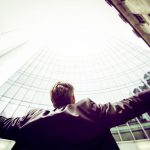 Mężczyzna w garniturze z rozpostartymi ramionami, a w tle wieżowce znikające w chmurach