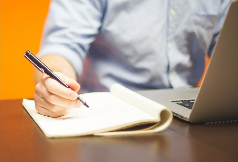 Fragment biurka, na biurku komputer i notatnik, mężczyzna w koszuli robi notatki